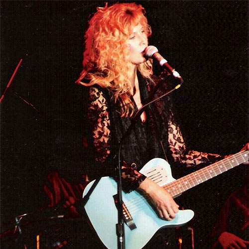 Gigi Dixon, with curly blonde hair, plays an electric guitar and sings into a microphone on stage, wearing a black lace outfit.