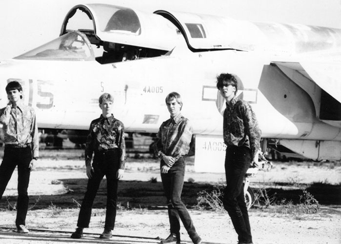 Four men stand in front of a military jet aircraft outdoors, all in patterned shirts and dark pants. The jet's canopy is open, with jagged rocks on the perimeter framing the dry, barren landscape.