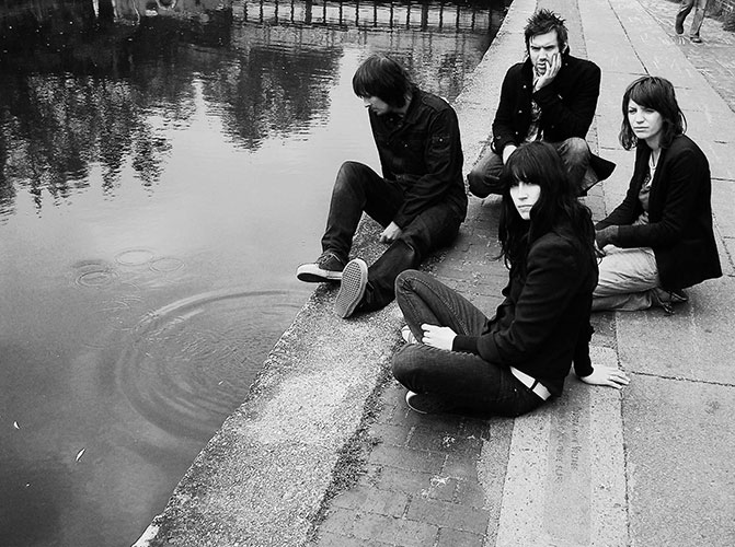 Four people sit and kneel by the edge of a body of water, looking contemplative. Trees and a railing are visible in the background. The image, reminiscent of a still from Scanners, is in black and white.