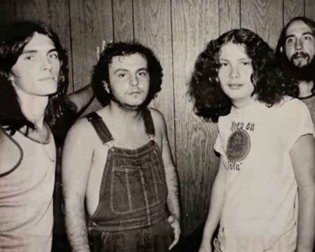 Four men with long hair stand in front of a wood-paneled wall; one wears overalls, the others wear casual shirts, capturing the laid-back Southern style of the Tennessee River Crooks.