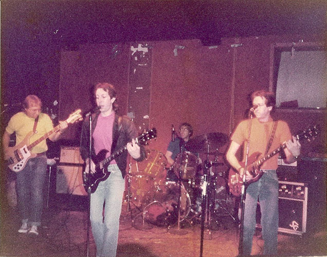 Four musicians perform on stage with guitars and drums in a dimly lit venue, embracing the calculated risk that comes with every live show.