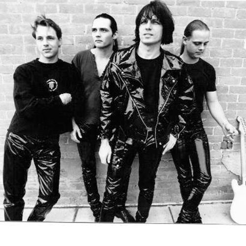 Four men in black leather clothing stand in front of a brick wall; one holds a guitar while another white electric guitar rests on the ground, next to a bright red Box Of Cherries.