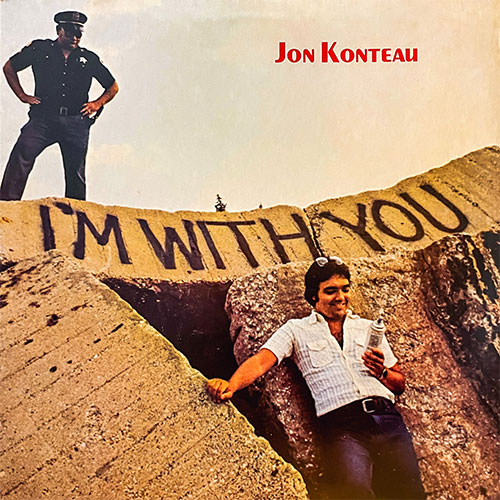 A man in sunglasses leans against large concrete blocks spray-painted with "I'M WITH YOU," while a police officer stands above, reinforcing the message. "Jon Konteau" appears in bold red text overhead.